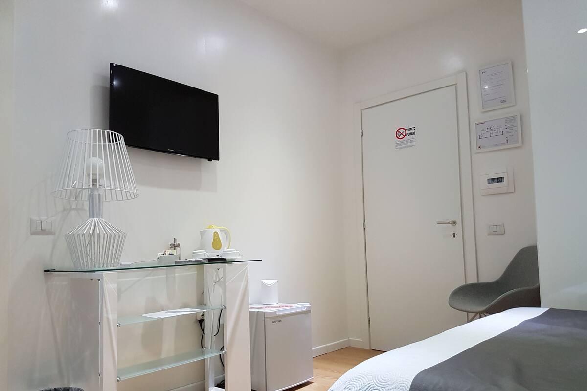 A bedroom with a bed, a TV mounted on the wall, a small kitchenette with a fridge, and a door with a 'no smoking' sign.