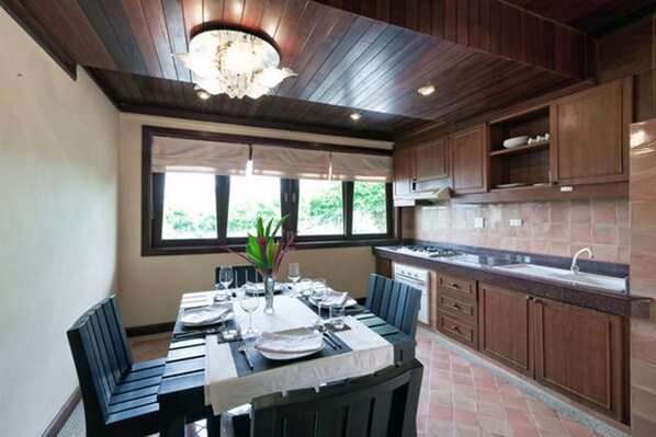 A modern kitchen with wooden cabinets, a dining area with a table set for four, and a large window providing natural light. - Villa H (Kamala)