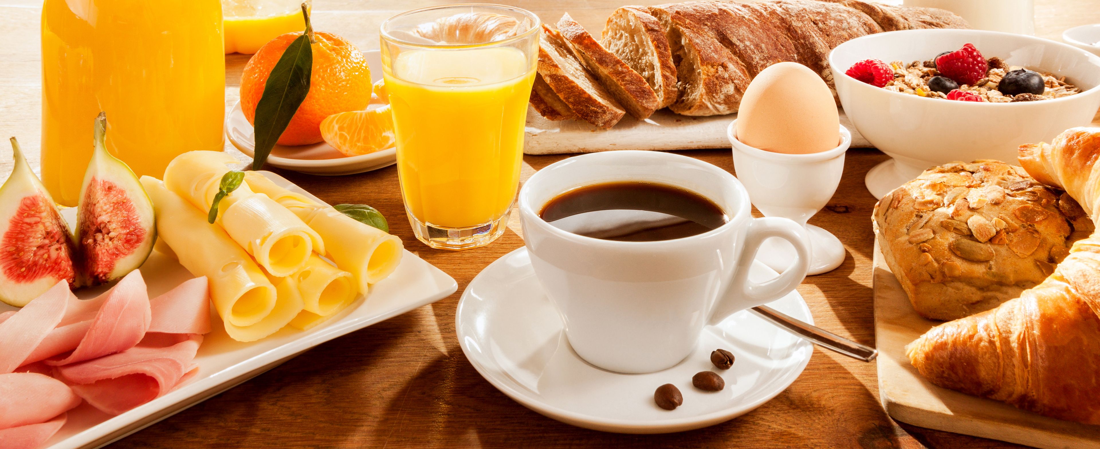 Une table de petit-déjeuner avec des croissants, un croissant coupé en deux, une assiette de jambon, de fromage et de figues, un verre de jus d’orange, une tasse de café et un bol de céréales aux fruits rouges.