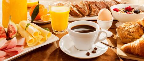 Une table de petit-déjeuner avec des croissants, un croissant coupé en deux, une assiette de jambon, de fromage et de figues, un verre de jus d’orange, une tasse de café et un bol de céréales aux fruits rouges.