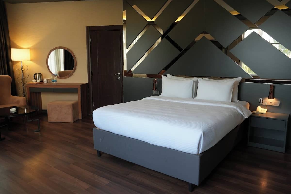 A modern hotel room with a large bed, a desk, a chair, a round mirror, and a unique geometric ceiling design.