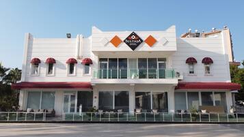 A two-story building with a sign reading "SULTASA HOTEL". There are multiple windows on the ground floor, a balcony on the second floor, and a red awning over the entrance.