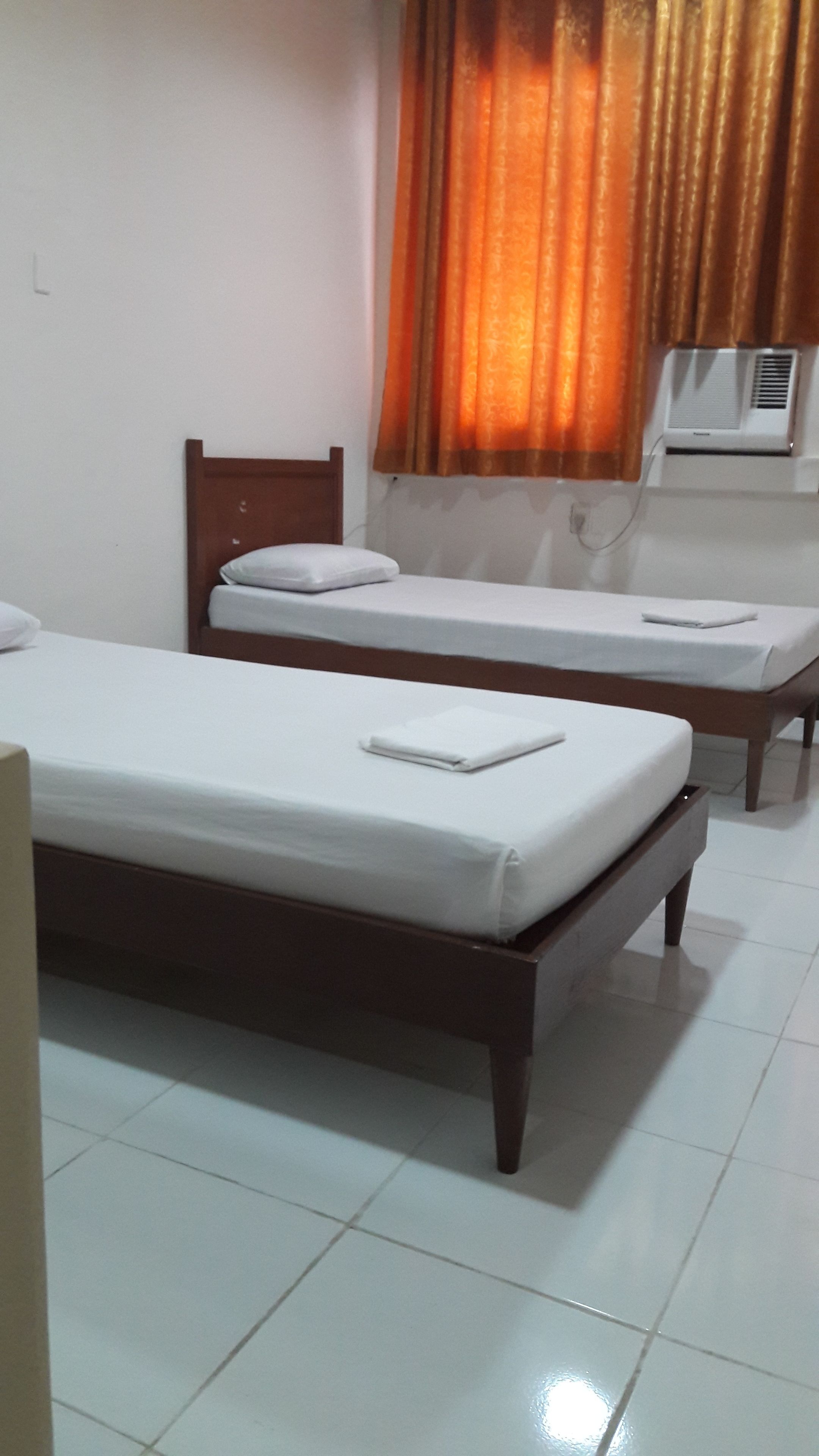 Two single beds with wooden frames and white linens, a window with an air conditioning unit, and a tiled floor.