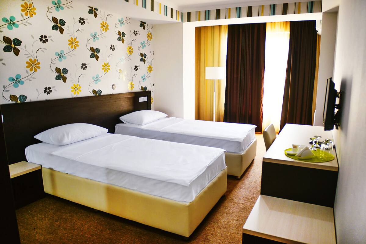 A hotel room with two single beds, a desk with a chair, a TV, and a window with curtains.
