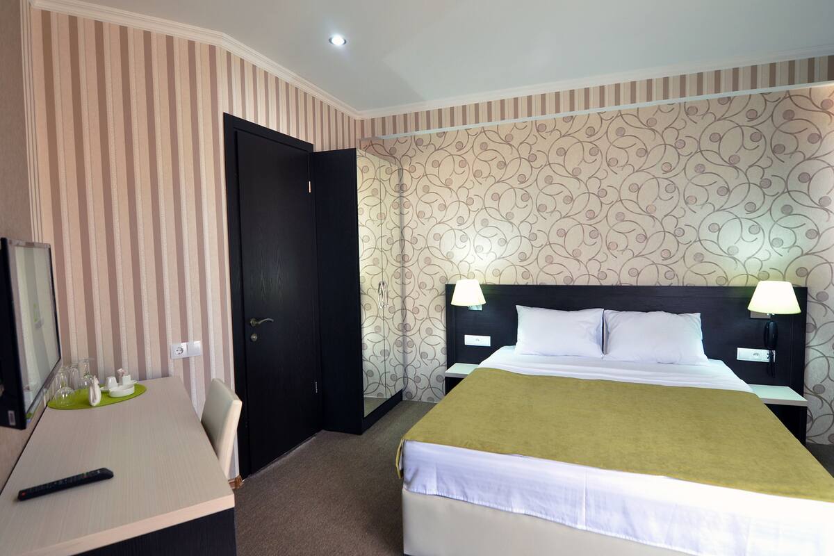 A hotel room with a bed, two bedside tables with lamps, a desk with a remote control, a chair, and a door.