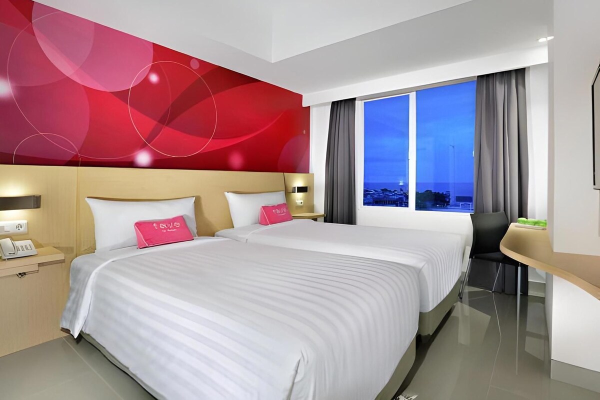 Habitación de hotel con dos camas, un escritorio y un ventanal con vistas a la ciudad.