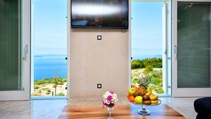A room with a large flat-screen TV mounted on the wall, a wooden table with a fruit bowl, and a vase of flowers. - View 4 You Apartments (Podstrana)