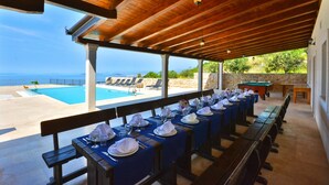 A covered outdoor dining area with tables set for a meal, overlooking a pool and the sea. - View 4 You Apartments (Podstrana)