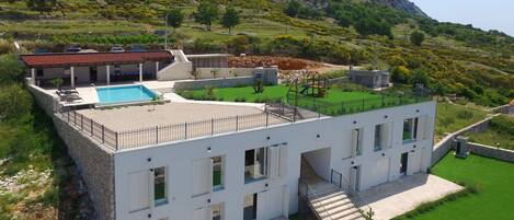 A modern villa with a swimming pool, a fenced garden, and a view of a mountainous landscape.