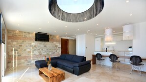 A modern living room with a stone wall, a sectional sofa, a wooden coffee table, and a kitchen area with bar stools. - View 4 You Apartments (Podstrana)