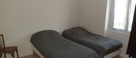 A small, single-room with two single beds, a wooden chair, and a window with curtains.