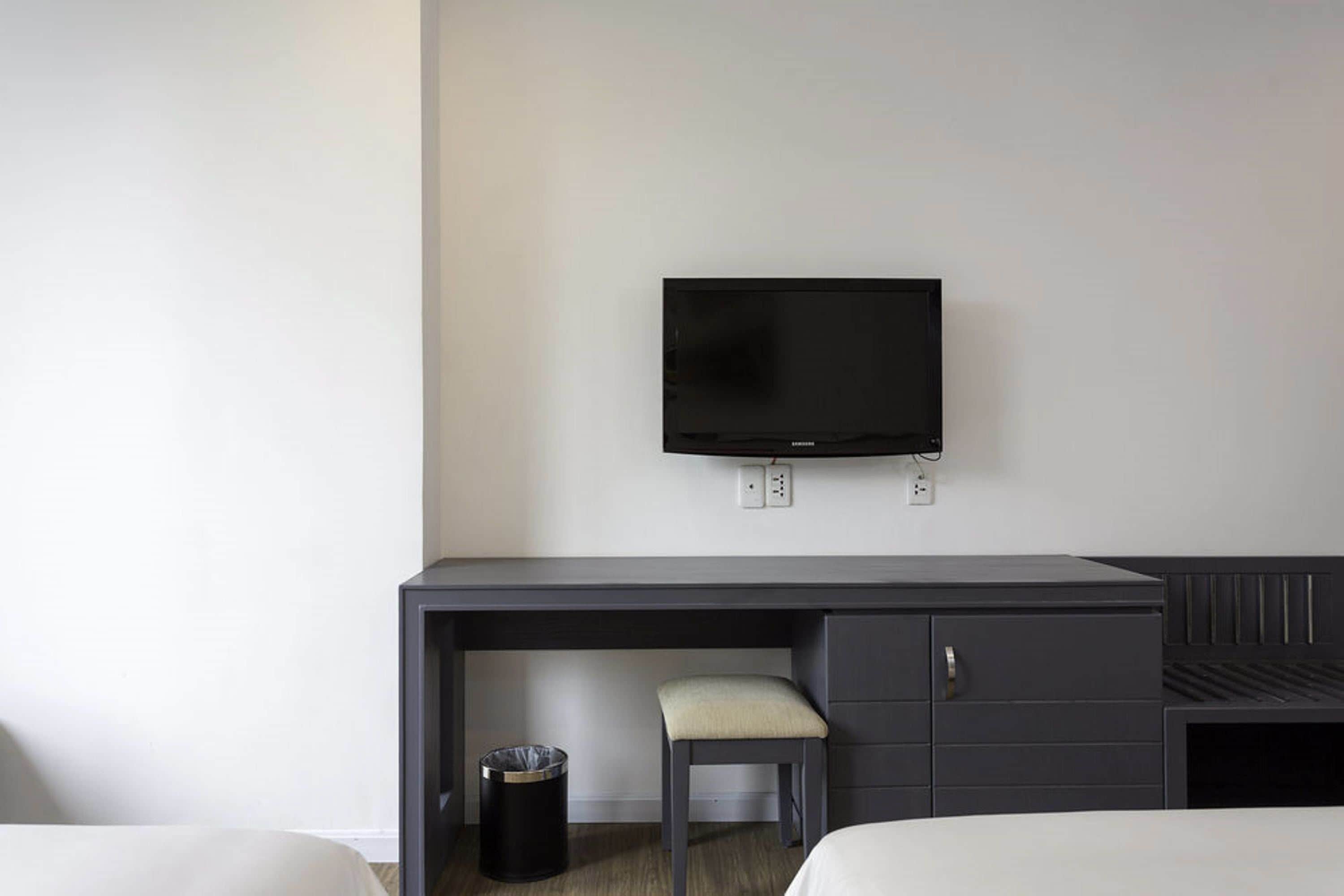 A hotel room with a bed, a desk with a chair, a television mounted on the wall, and a trash bin.