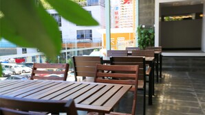 Un restaurant avec des tables et des chaises en bois, une vue sur un parking et un bâtiment sur lequel on peut lire une enseigne dans une langue étrangère.