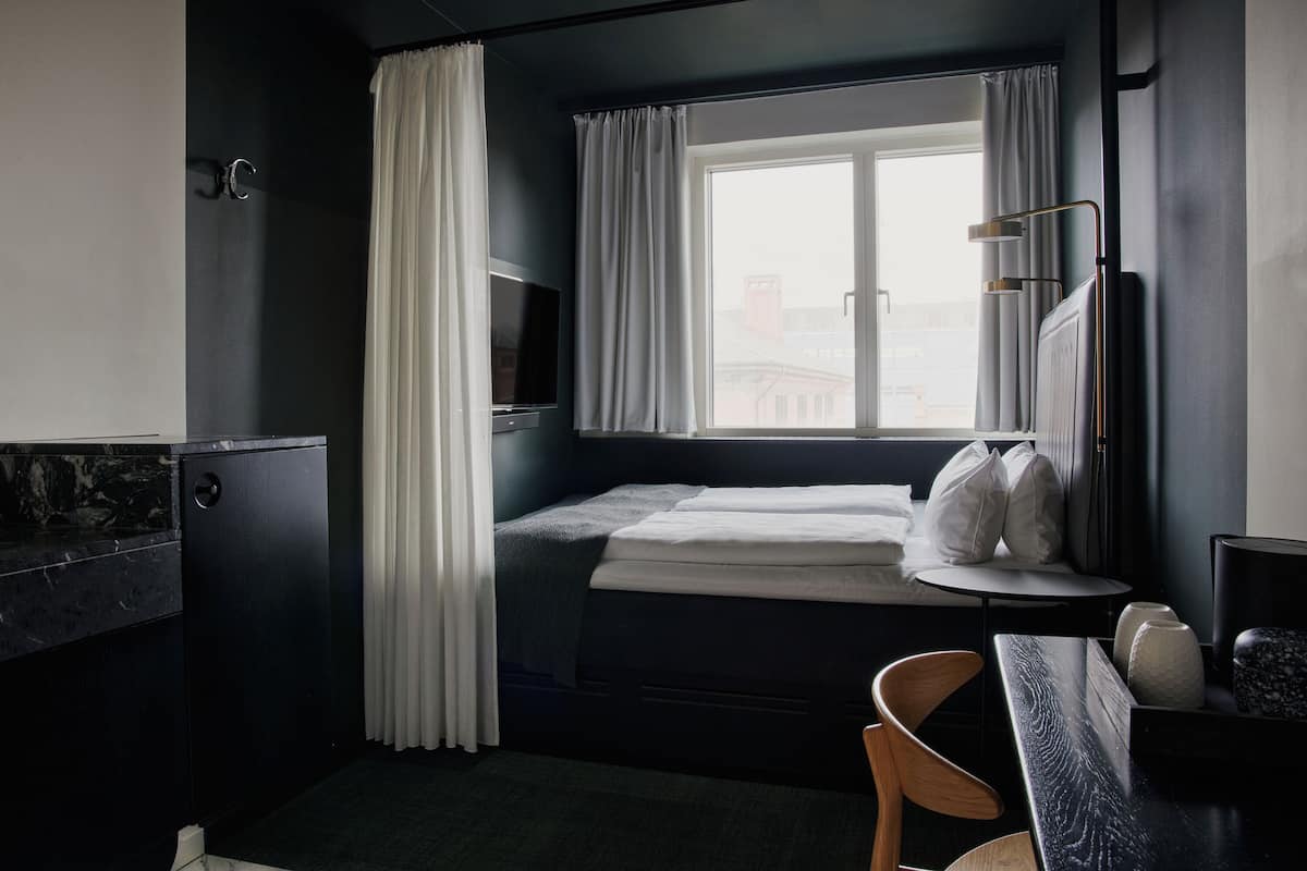 A compact hotel room with a bed, a desk, a chair, a TV, and a window with curtains.