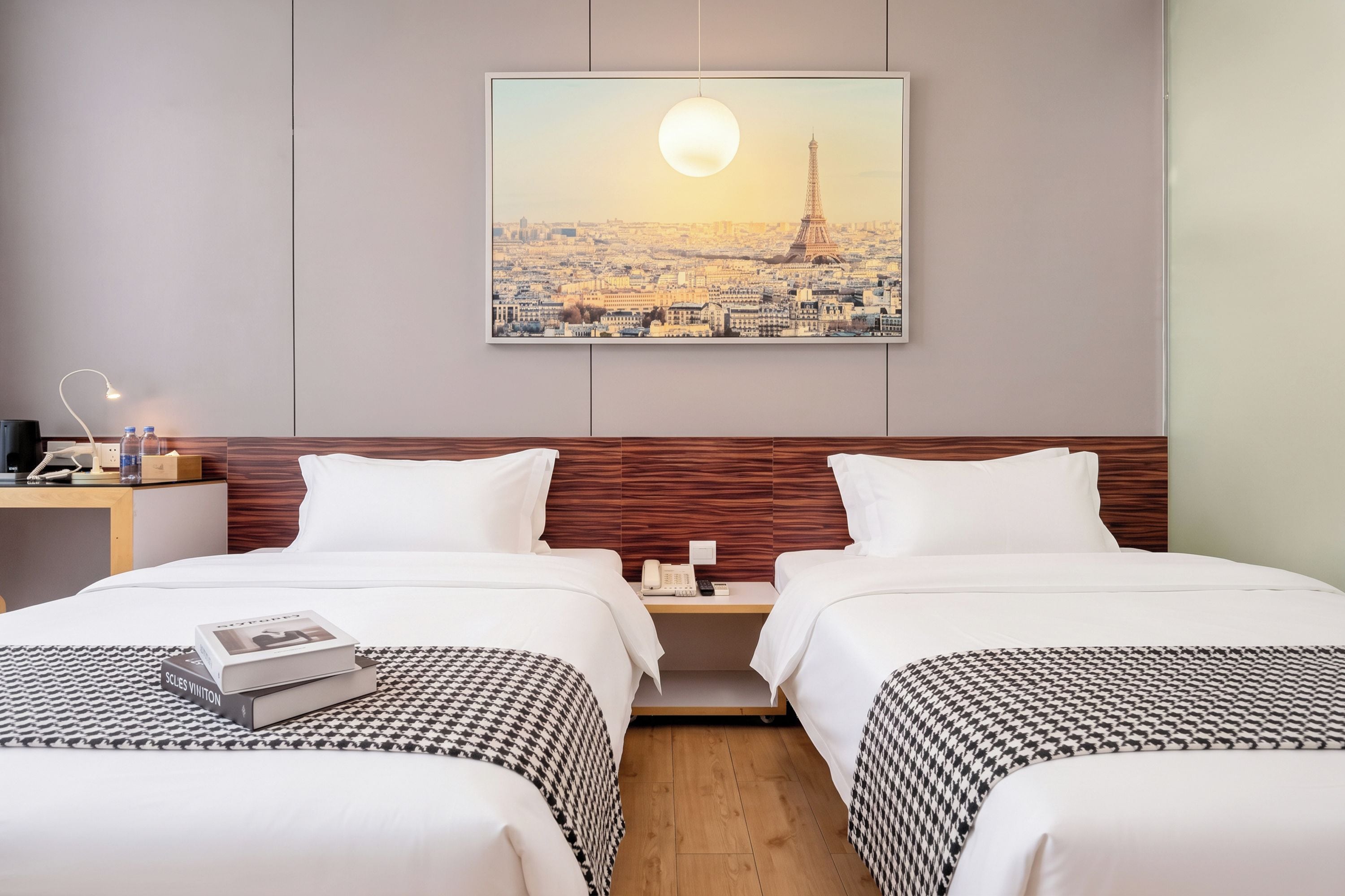 A hotel room with two beds, a nightstand with a lamp, a phone, and a framed picture of the Eiffel Tower on the wall.