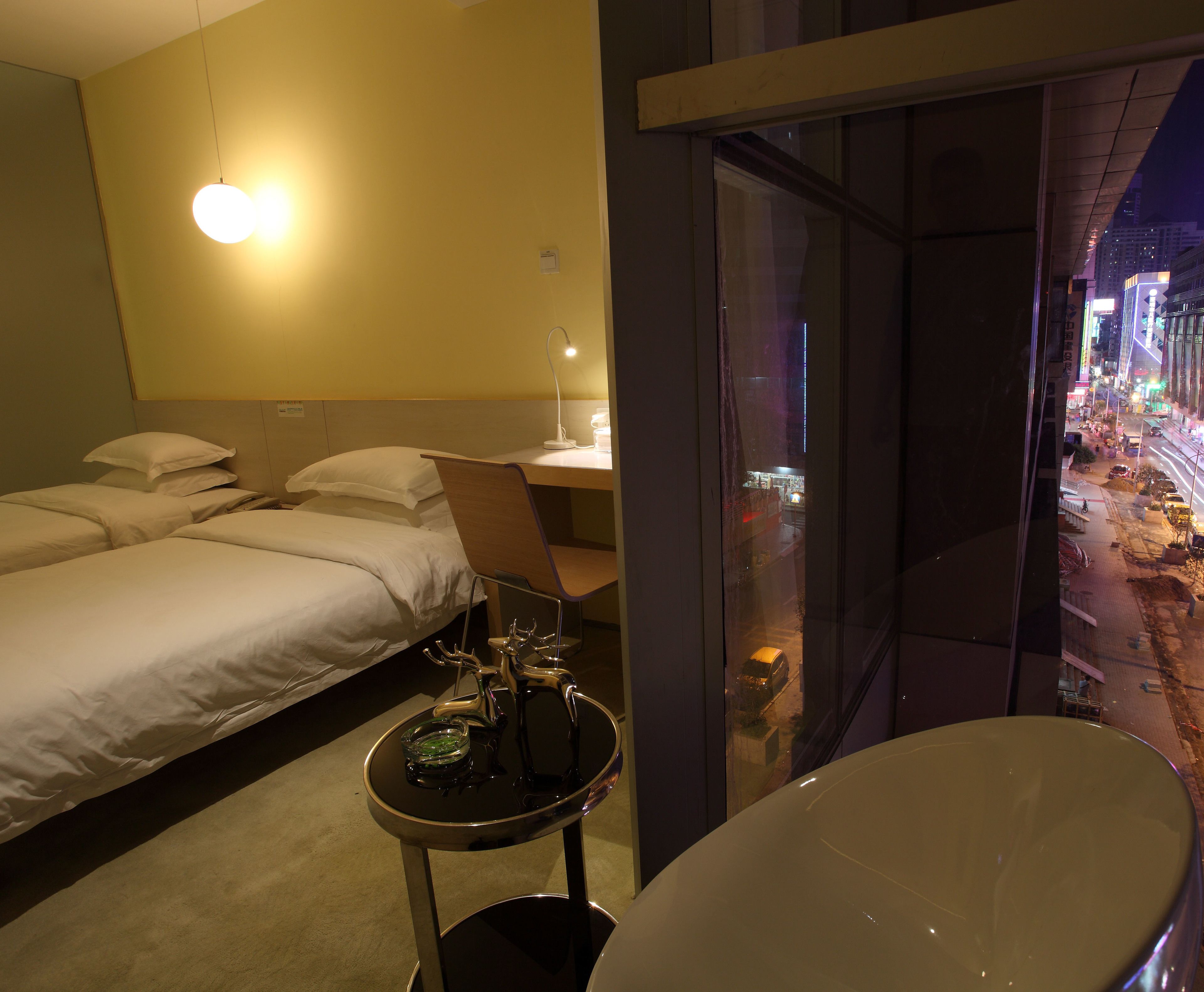 A hotel room with two beds, a desk, a chair, a small table with a glass, and a large window offering a city view.