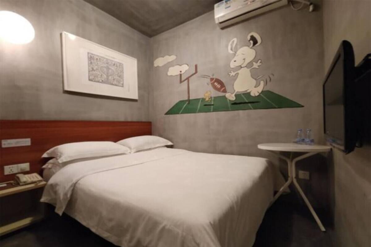 A hotel room with a bed, a small bedside table with a lamp, a wall-mounted air conditioner, and a wall decoration of a cartoon character playing football.