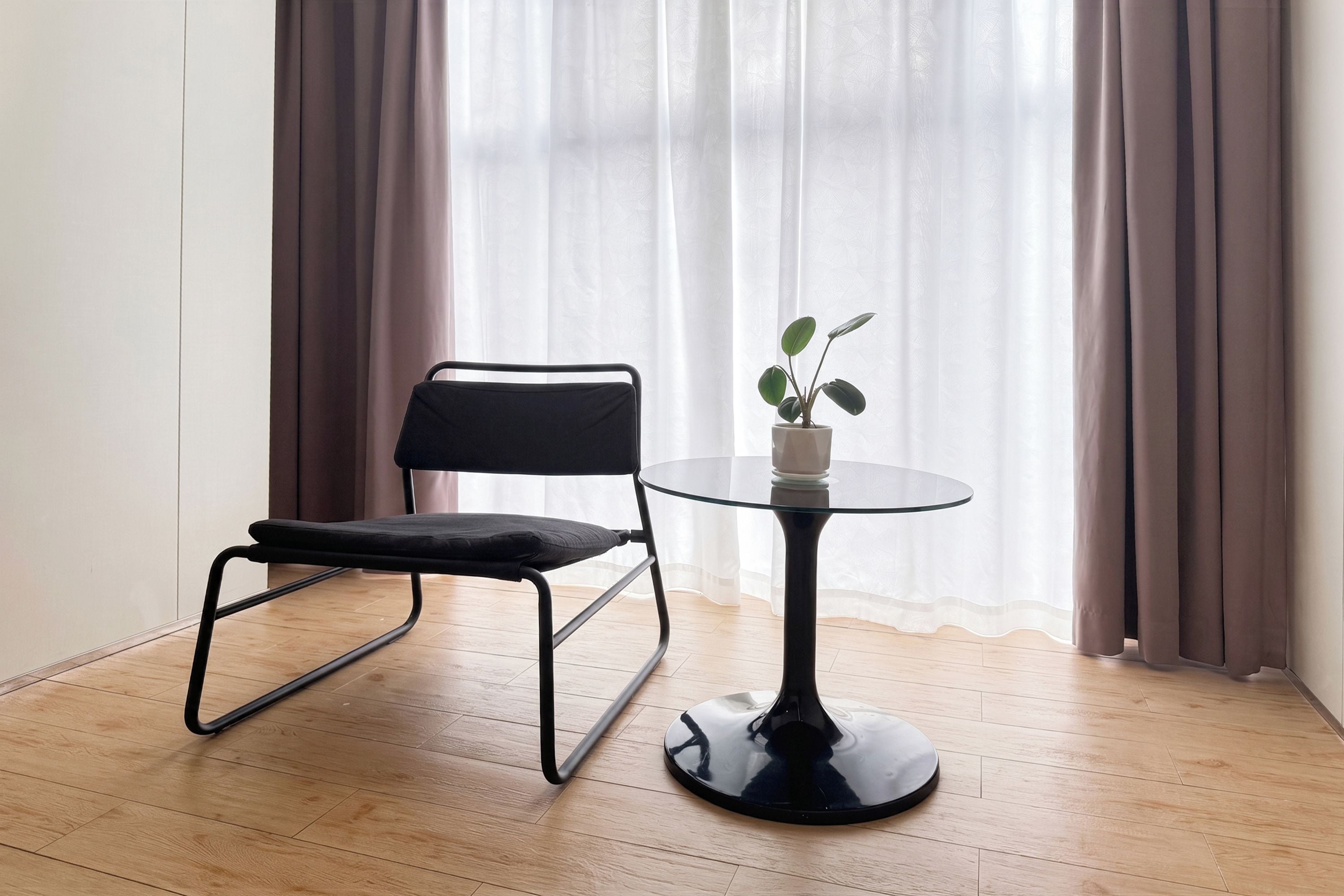 Una habitación con una silla negra, una mesita con una planta y una ventana con cortinas.