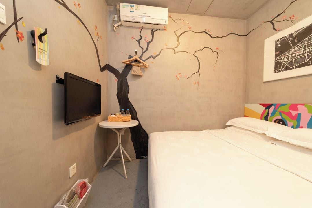 Quarto de hotel com cama, televisão presa à parede, mesa redonda pequena e decoração na parede que se assemelha a galhos de árvore.