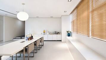 A modern kitchen with a long dining table, white cabinets, and a large window with blinds.
