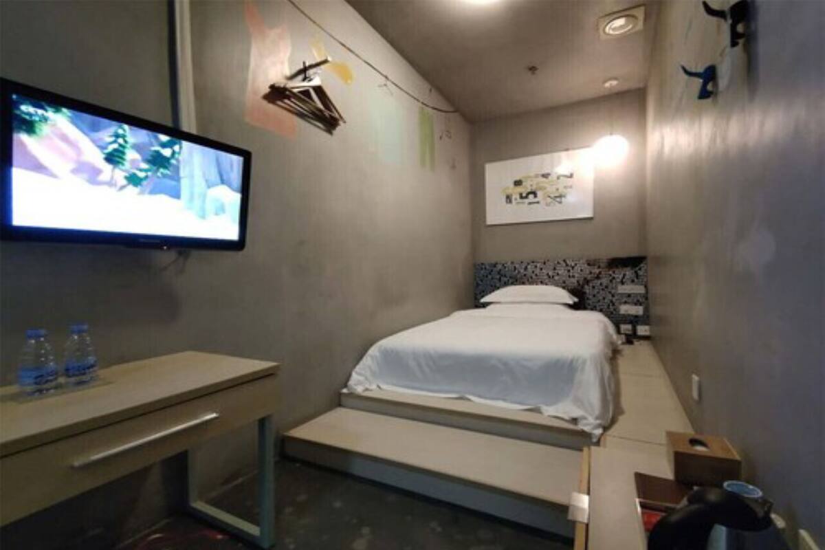 A small, modern hotel room with a bed, a desk, a TV, and a wall-mounted light fixture.