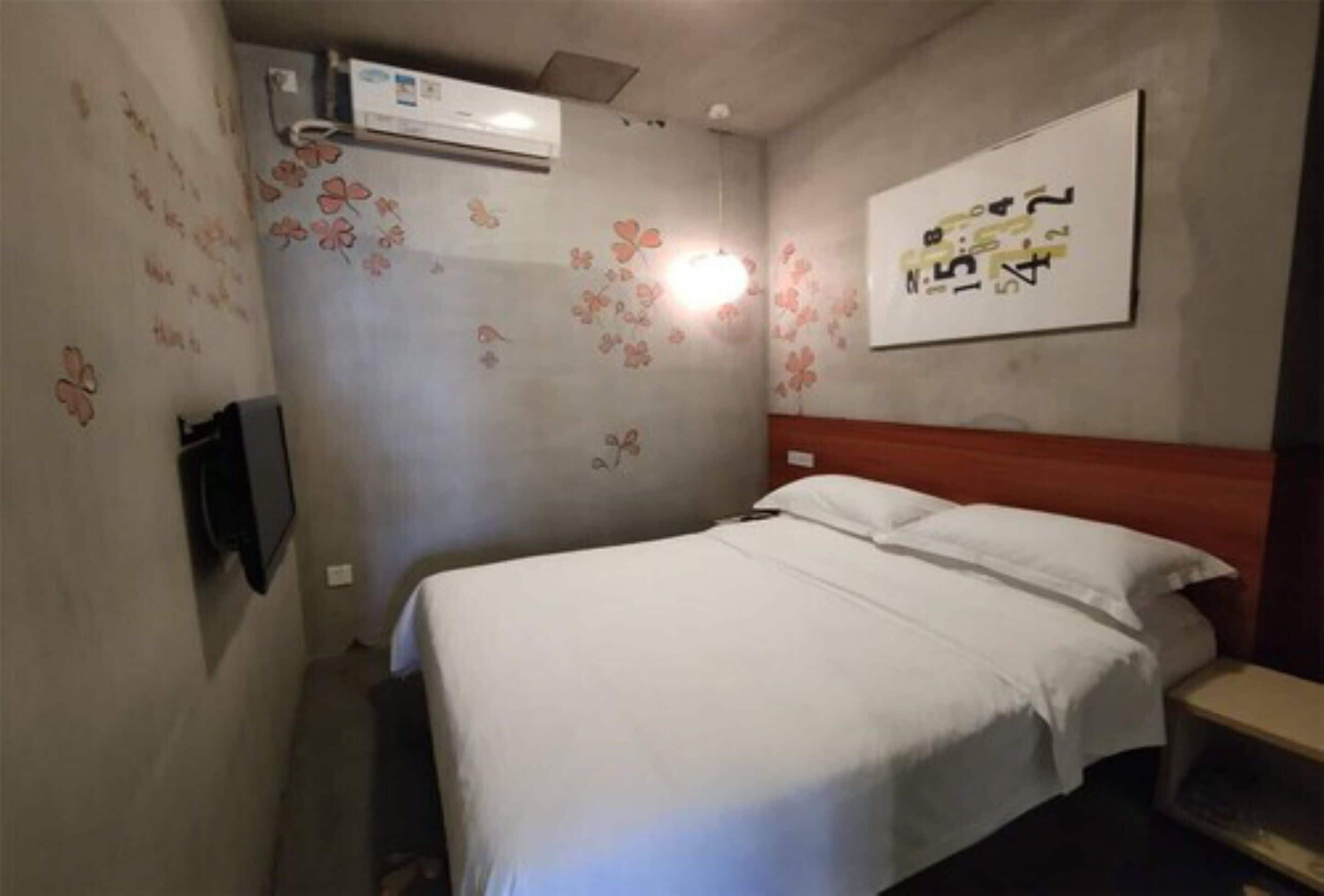 Une petite chambre avec un lit simple, une tête de lit en bois, un téléviseur fixé au mur et des motifs floraux décoratifs sur le mur.