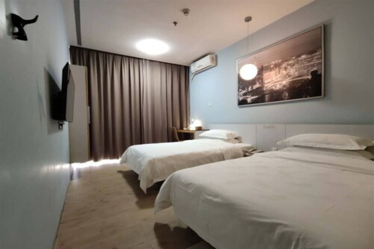 A hotel room with two beds, a flat-screen TV mounted on the wall, a nightstand with a lamp, and a large window with curtains.