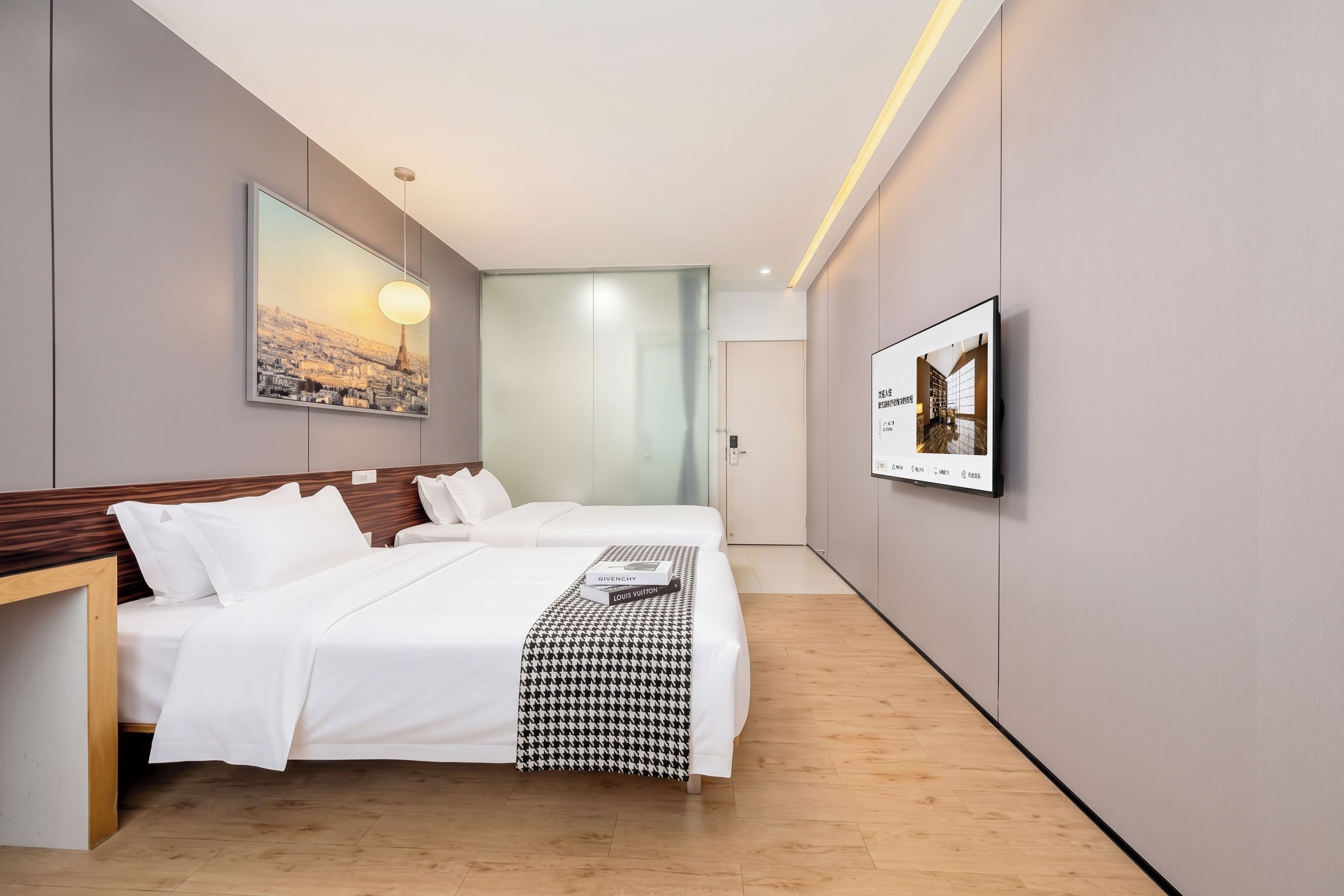 Habitación de hotel con dos camas, cabecera de madera, manta a cuadros de piedritas, pintura de un paisaje urbano, un cuadro enmarco y un televisor de pantalla plana.