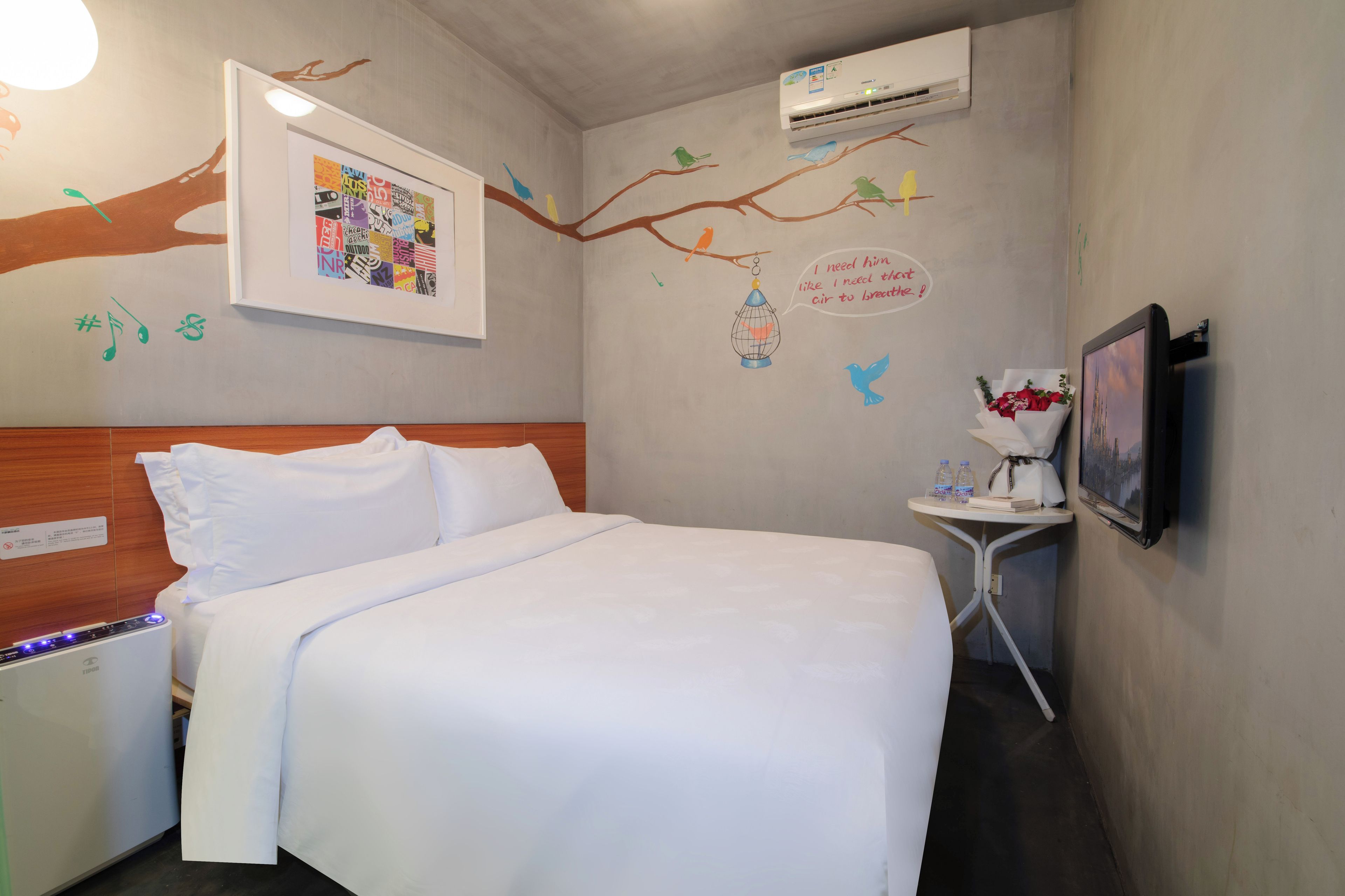 A hotel room with a bed, a small table with a vase of flowers, an air conditioner, and wall decorations including a framed picture and bird illustrations.