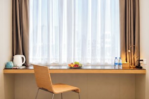A hotel room with a wooden desk, a chair, a window with sheer curtains, a fruit bowl, a kettle, and two water bottles.