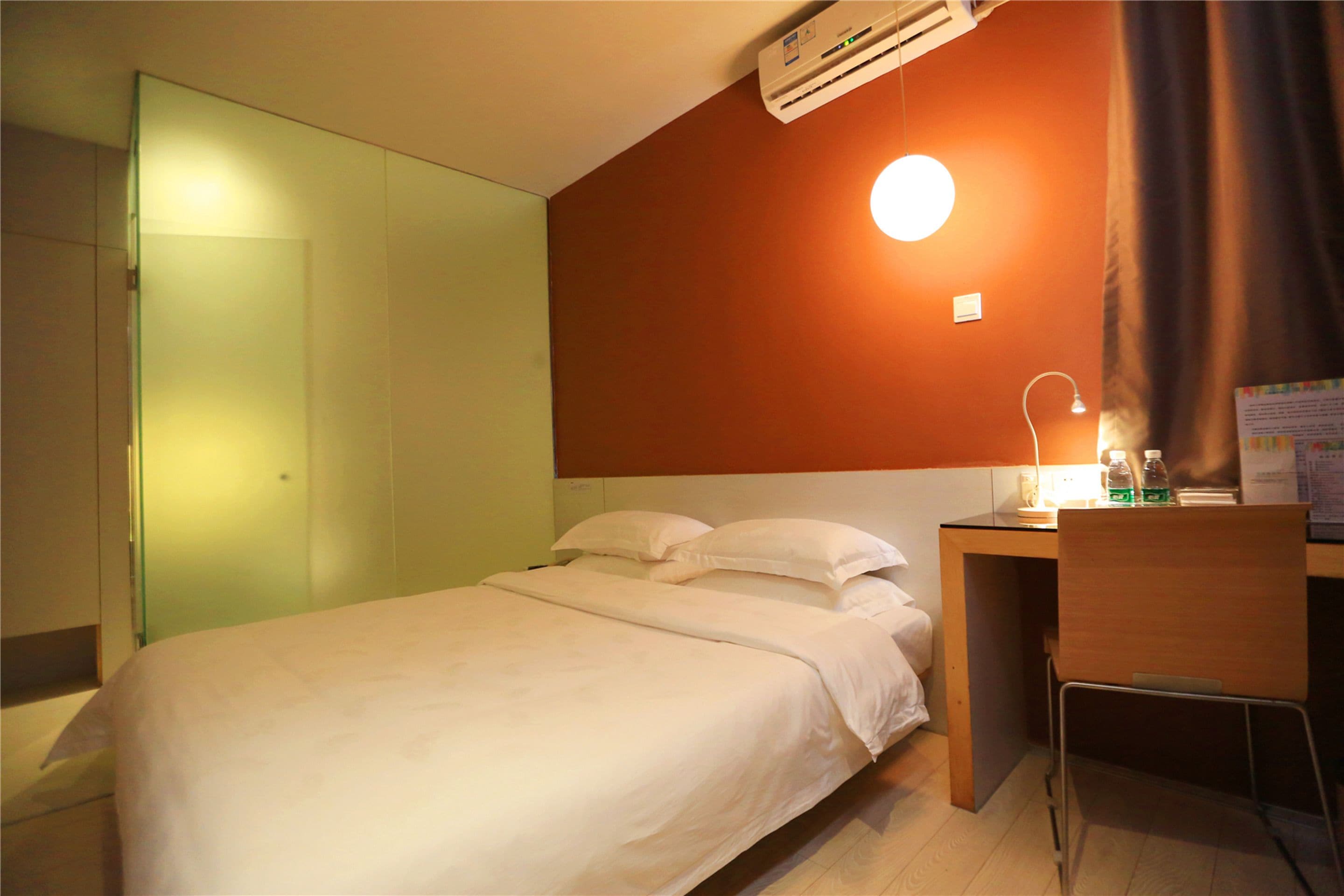 A hotel room with a bed, a desk, a chair, a wall-mounted air conditioner, a bedside lamp, and a wall-mounted light.