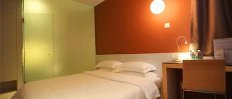 A hotel room with a bed, a desk, a chair, a wall-mounted air conditioner, a bedside lamp, and a wall-mounted light.