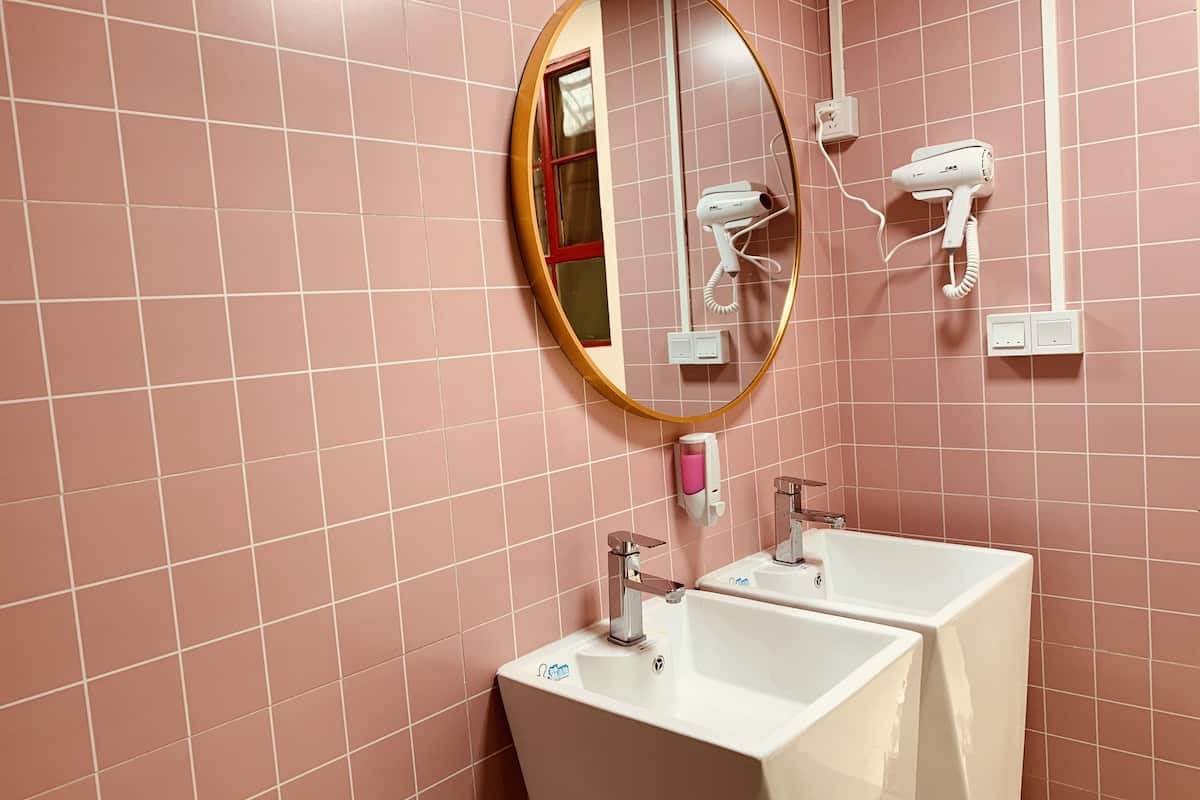 Un baño con paredes de azulejos rosados, un espejo redondo, dos lavamanos, un secador de pelo y un dispensador de jabón.