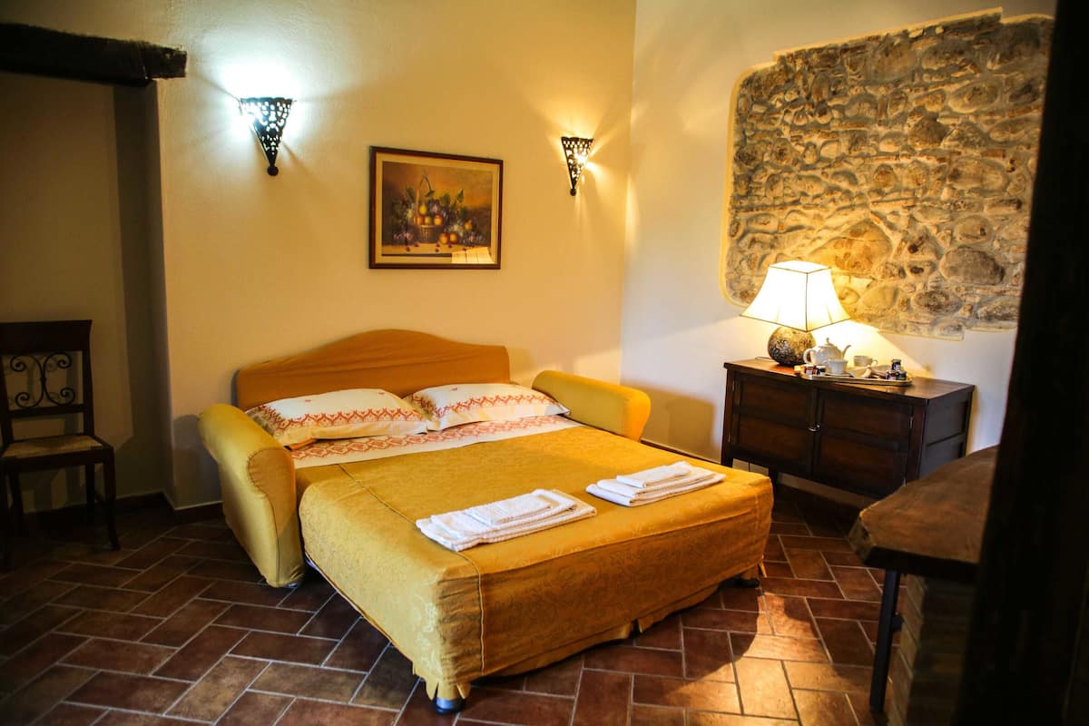 Une pièce avec un lit, une table de chevet avec une lampe, un tableau au mur et un mur en pierre.