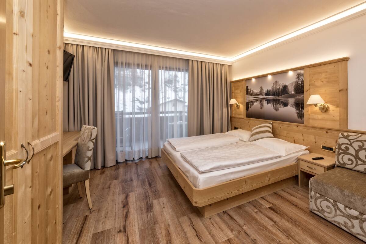 Une chambre d’hôtel avec un lit en bois, un canapé, un bureau et une grande fenêtre avec des rideaux.