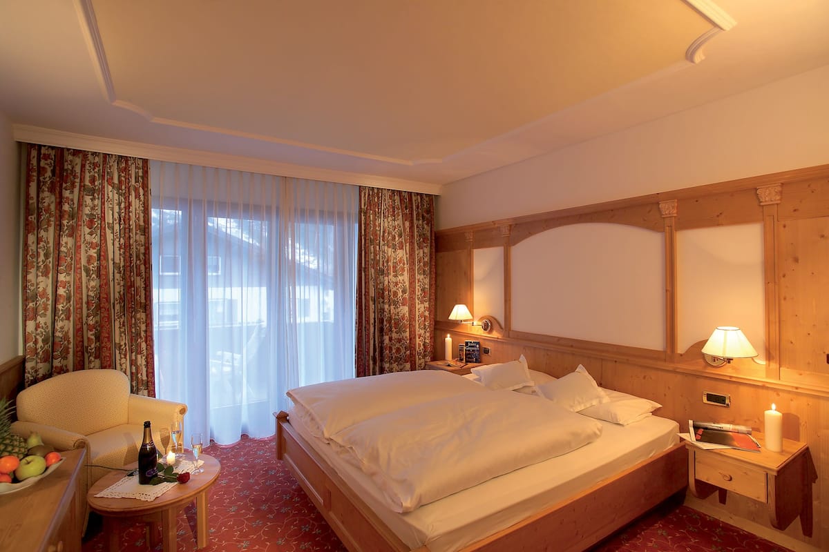 Une chambre d’hôtel avec un grand lit, une table de chevet en bois avec une lampe, une chaise, une petite table avec des fruits et une bouteille, et une fenêtre avec des rideaux.