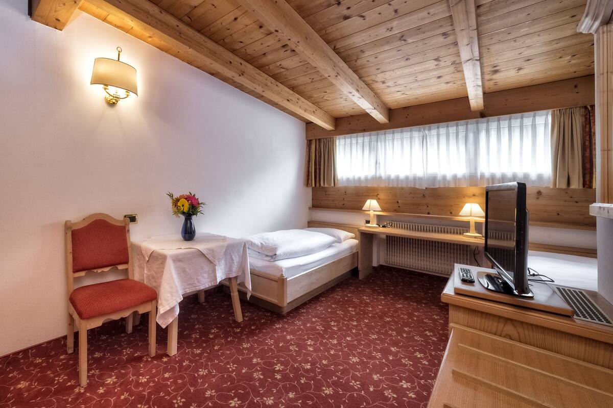 Une chambre d’hôtel avec un plafond en bois, un lit, un bureau avec un ordinateur, une chaise, une petite table avec un vase de fleurs et une fenêtre avec des rideaux.