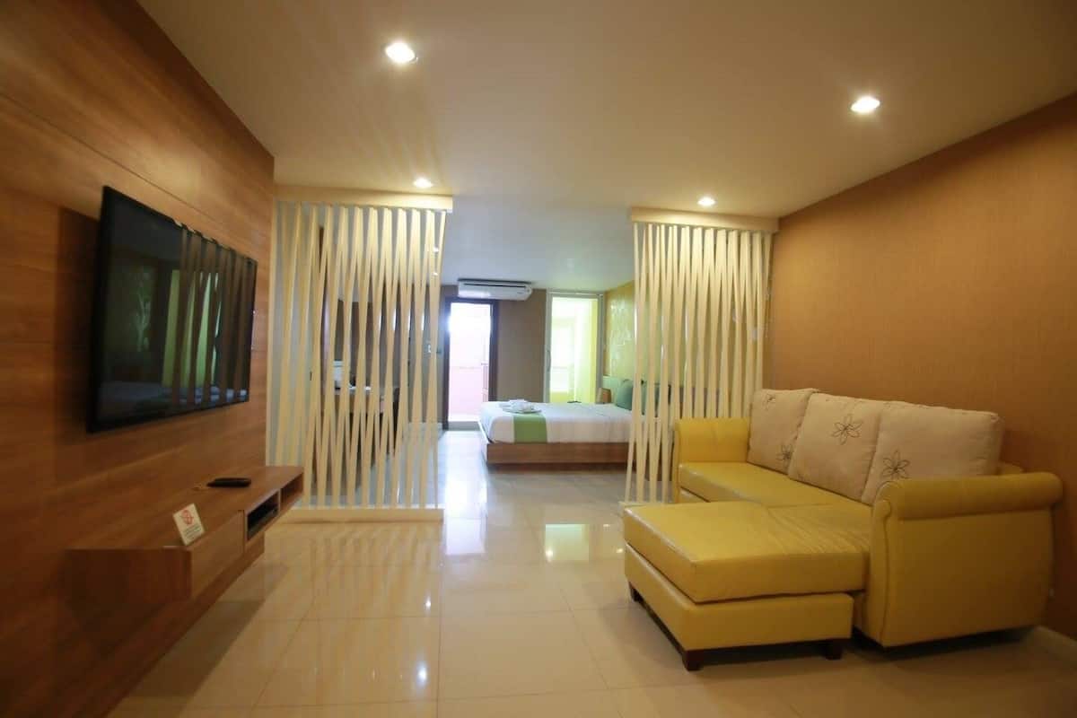 A modern hotel room with a wooden wall, a flat-screen TV, a yellow sofa, and a bed with a green bedspread.