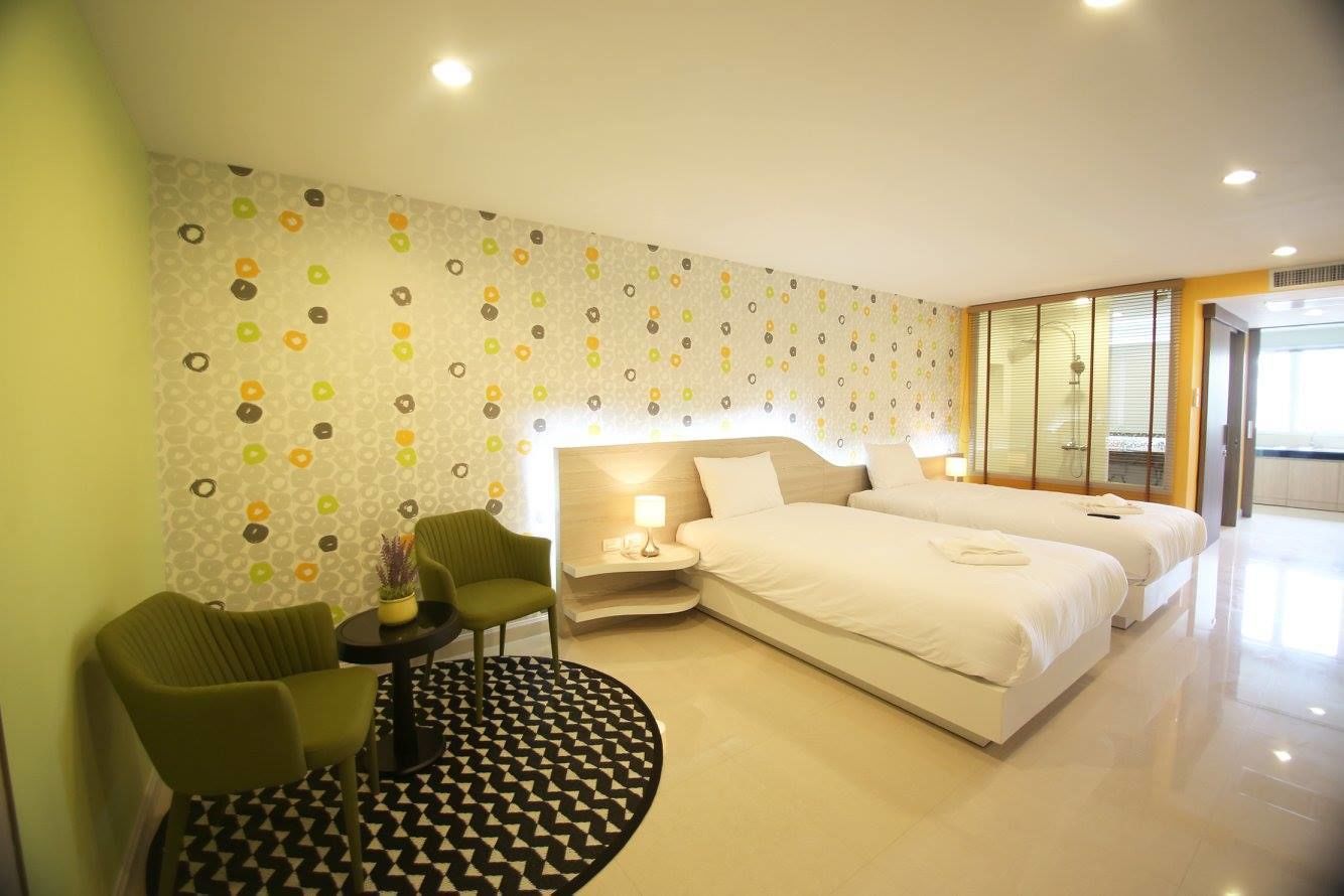 A hotel room with a large bed, two bedside tables, a green armchair, a small round table with a lamp, and a patterned wall.