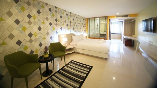 Quarto de hotel moderno com cama, área de estar com cadeiras, televisão e banheiro visível através de uma porta de vidro.