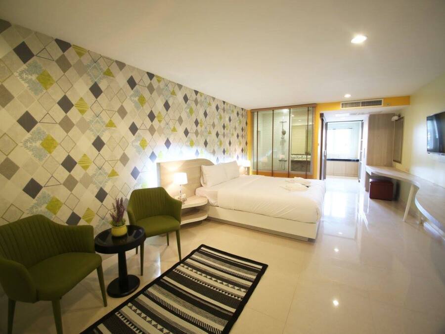Quarto de hotel moderno com cama, área de estar com cadeiras, televisão e banheiro visível através de uma porta de vidro.