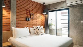 Habitació d'hotel moderna amb paret de maó, un llit gran amb roba blanca i una porta corredissa de vidre que dona a un balcó amb plantes.