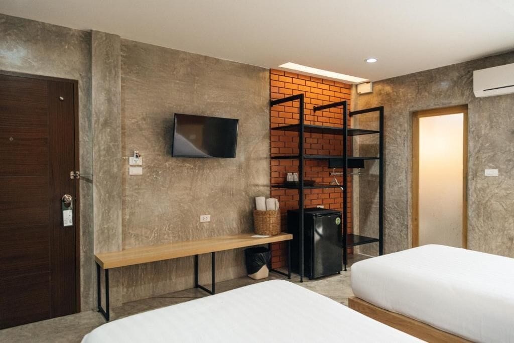 A modern hotel room with a brick accent wall, a flat-screen TV mounted above a wooden bench, a black refrigerator, and two beds.
