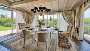 A dining room with a large table set for six, surrounded by wicker chairs, a chandelier, and large windows offering a view of greenery. - Villa Dora (San Gimignano)