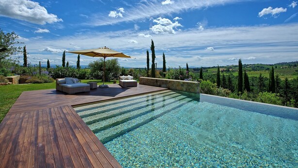 A poolside area with a wooden deck, two sofas, and an umbrella. - Villa Dora (San Gimignano)