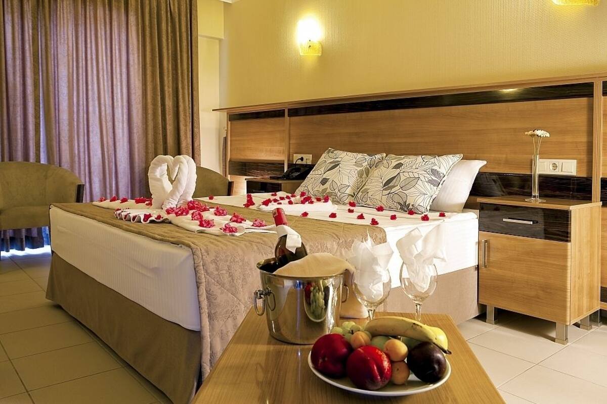A hotel room with a bed, a bedside table with a bottle of champagne, a fruit bowl, and a vase with flowers.