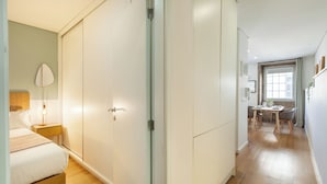 Un dormitorio con cama, mesita de noche y lámpara; un pasillo que conduce a un comedor con mesa y sillas; y una cocina con ventana y encimera.