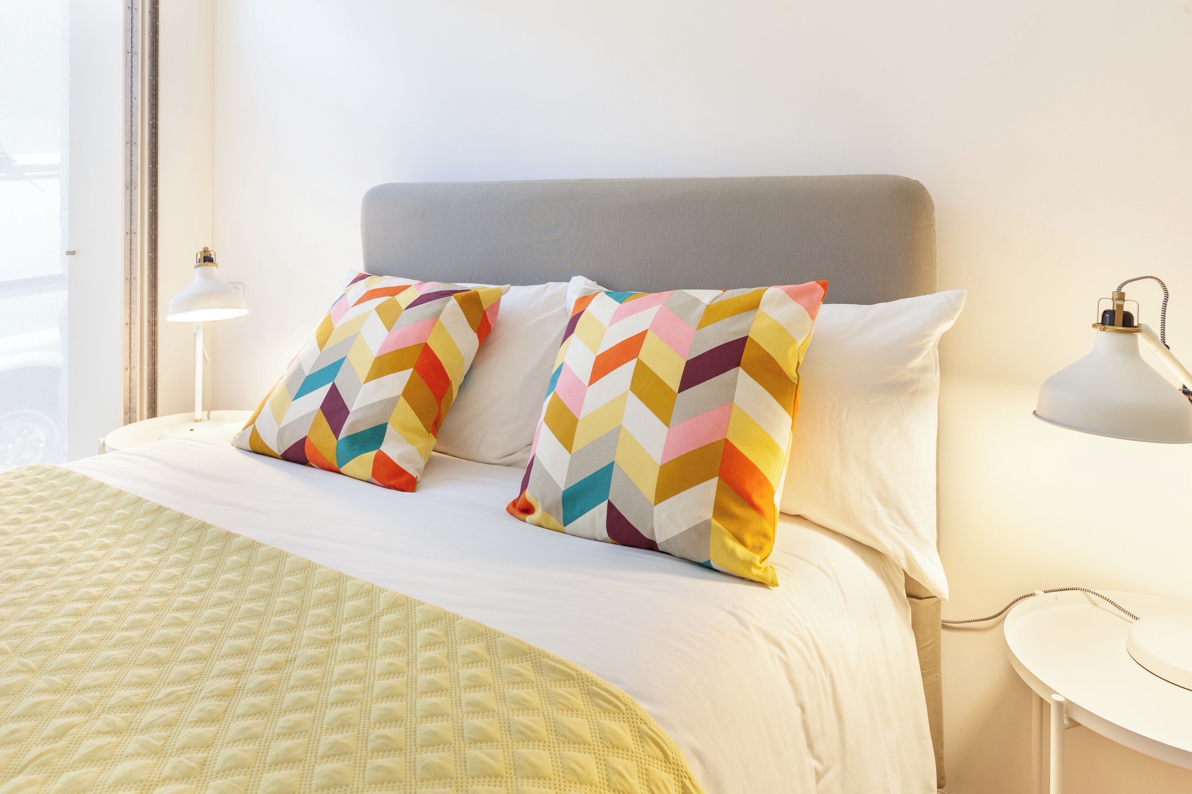 A neatly made bed with a quilted yellow blanket, two pillows with a multicolored chevron pattern, and a gray headboard.