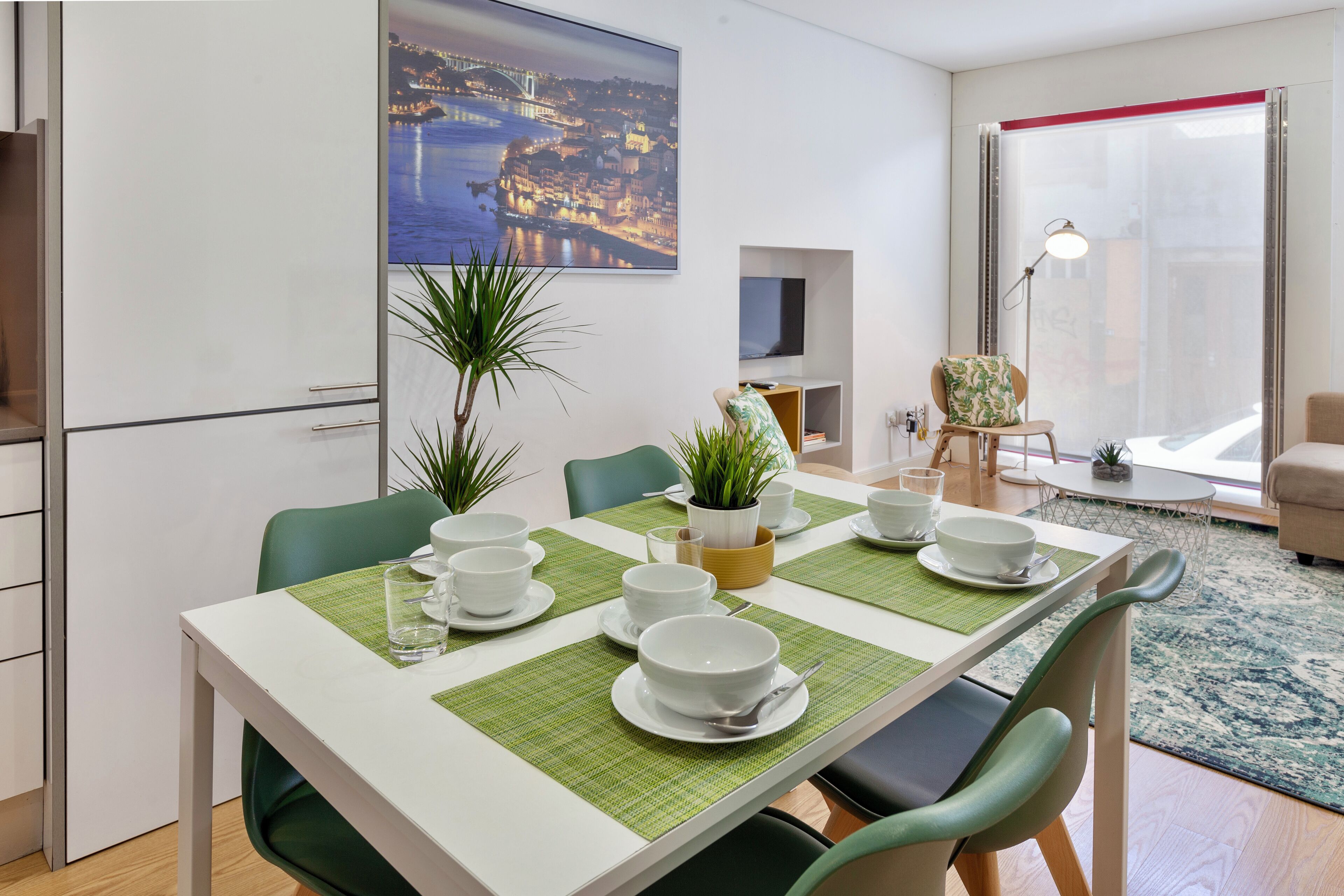 A dining area with a table set for four, green chairs, a potted plant, and a cityscape painting on the wall.