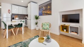 Una sala moderna con un televisor de pantalla plana, una zona de comedor con una mesa blanca y sillas verdes, y una cocina con armarios blancos.